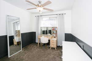 Office space with a wainscoted wall, dark colored carpet, a ceiling fan, and a decorative wall
