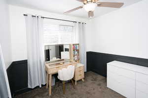 Office featuring a wainscoted wall, dark carpet, ceiling fan, and a decorative wall