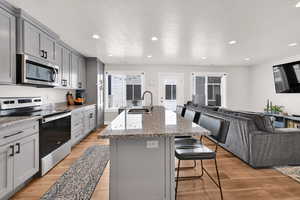 Kitchen featuring gray cabinetry, stainless steel appliances, open floor plan, light stone countertops, and a kitchen island with sink
