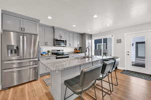 Kitchen with gray cabinetry, stainless steel appliances, light stone counters, light wood finished floors, and recessed lighting