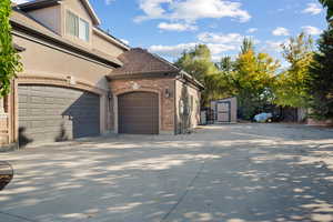 View of side of home featuring brick siding, a storage shed, stucco siding, driveway, and roof with shingles