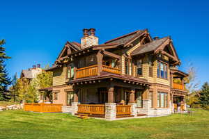 Rear view of house featuring stone siding, a chimney, a balcony, and a lawn