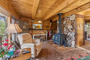 Sitting room with wood walls, carpet floors, a wood stove, and a wooden ceiling with exposed beams