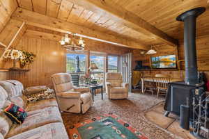 Carpeted living room featuring a wood stove, wooden walls, hanging lights, and a wooden ceiling with exposed beams
