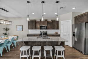 Kitchen featuring stainless steel appliances, light stone counters, dark wood finish cabinets, a center island with sink, and a textured ceiling