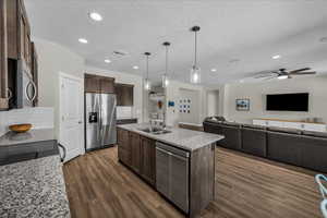 Kitchen featuring open floor plan, light stone counters, dark wood finish cabinets, stainless steel appliances, and a kitchen island with sink