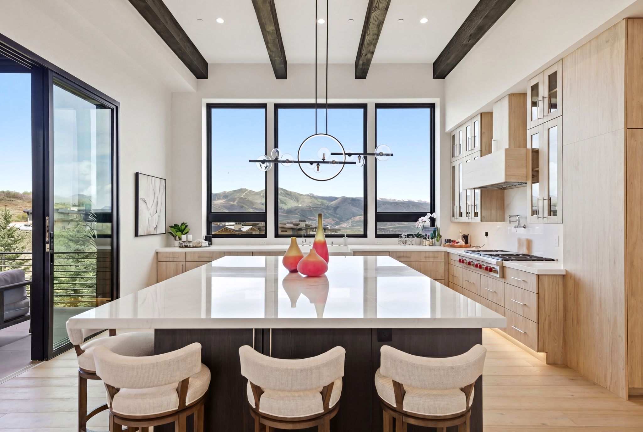 Gourmet kitchen featuring a large central island with seating, panoramic windows, exposed ceiling beams, light wood-finish cabinetry, and stainless steel appliances