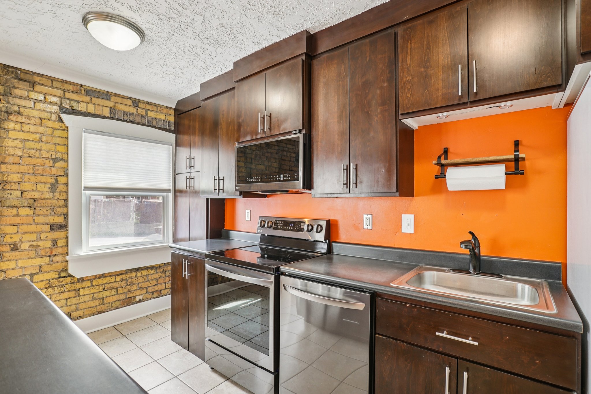 Kitchen featuring dark wood finish cabinetry, dark countertops, stainless steel appliances, brick wall, and light tile patterned flooring