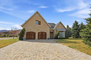 French country style house with a front yard, stone siding, decorative driveway, and an attached garage