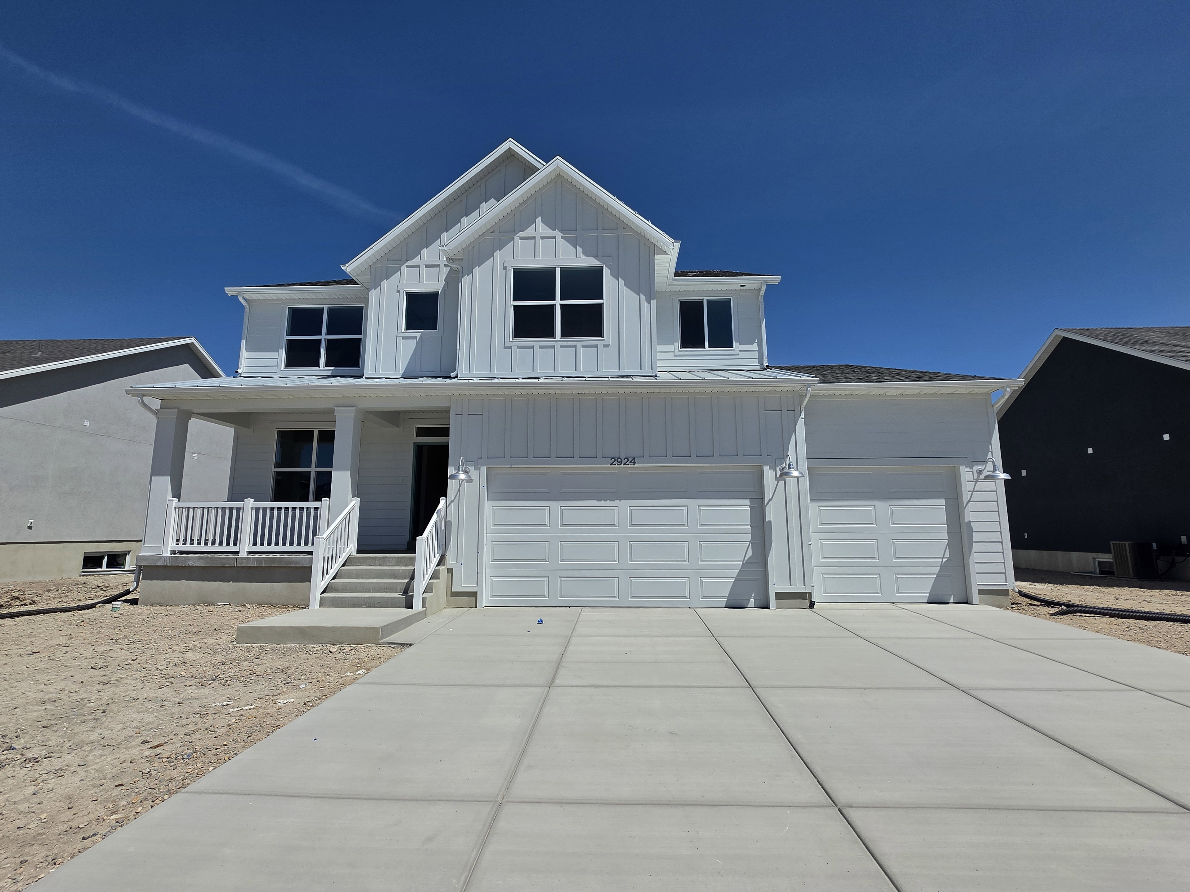 Two-story residence featuring white board and batten siding, a covered porch with white railings, a two-car garage with panel doors, and a concrete driveway