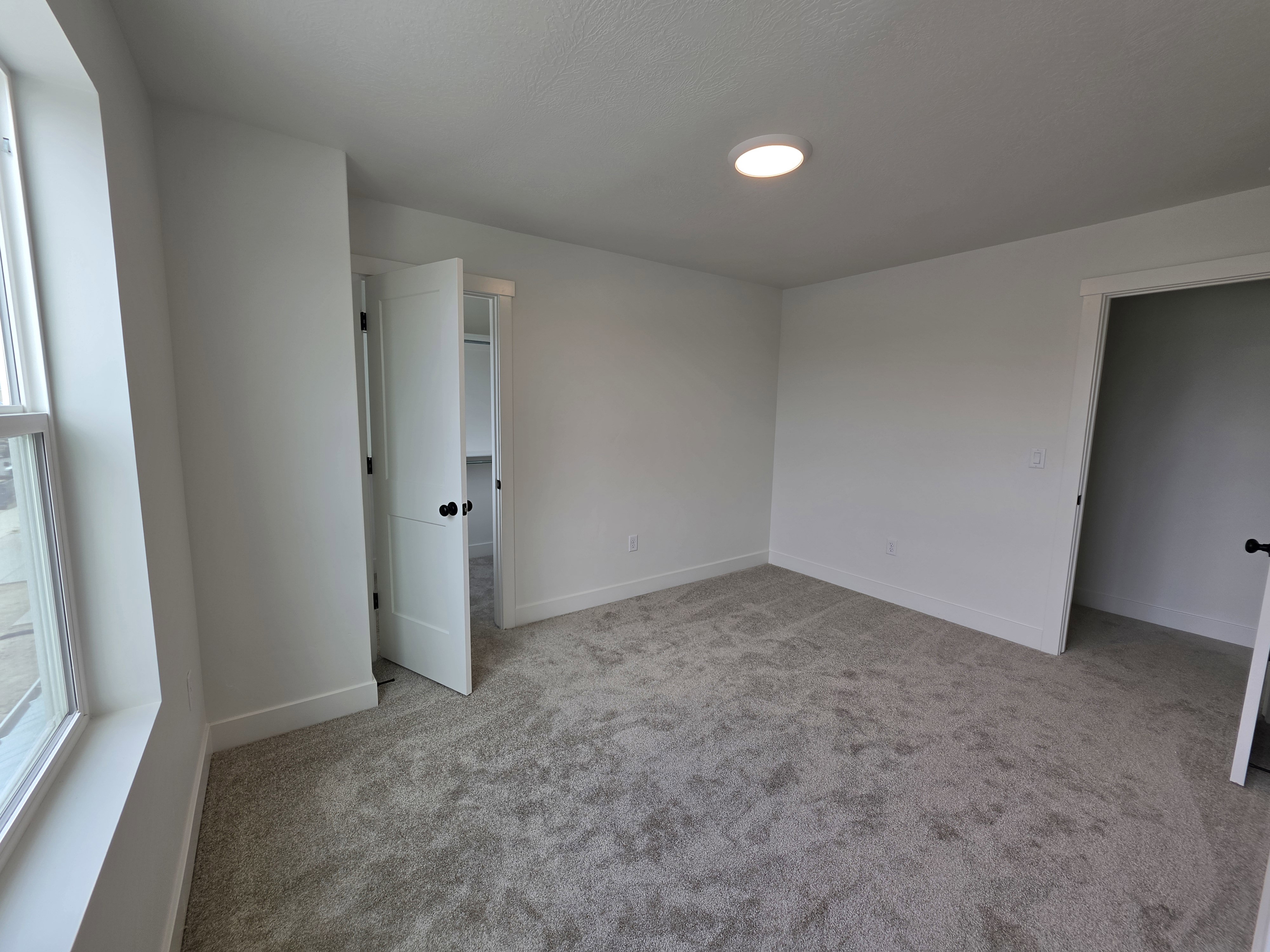 Spacious room featuring neutral carpeting, white walls, a large window, a flush-mount ceiling light, and white interior doors with dark hardware