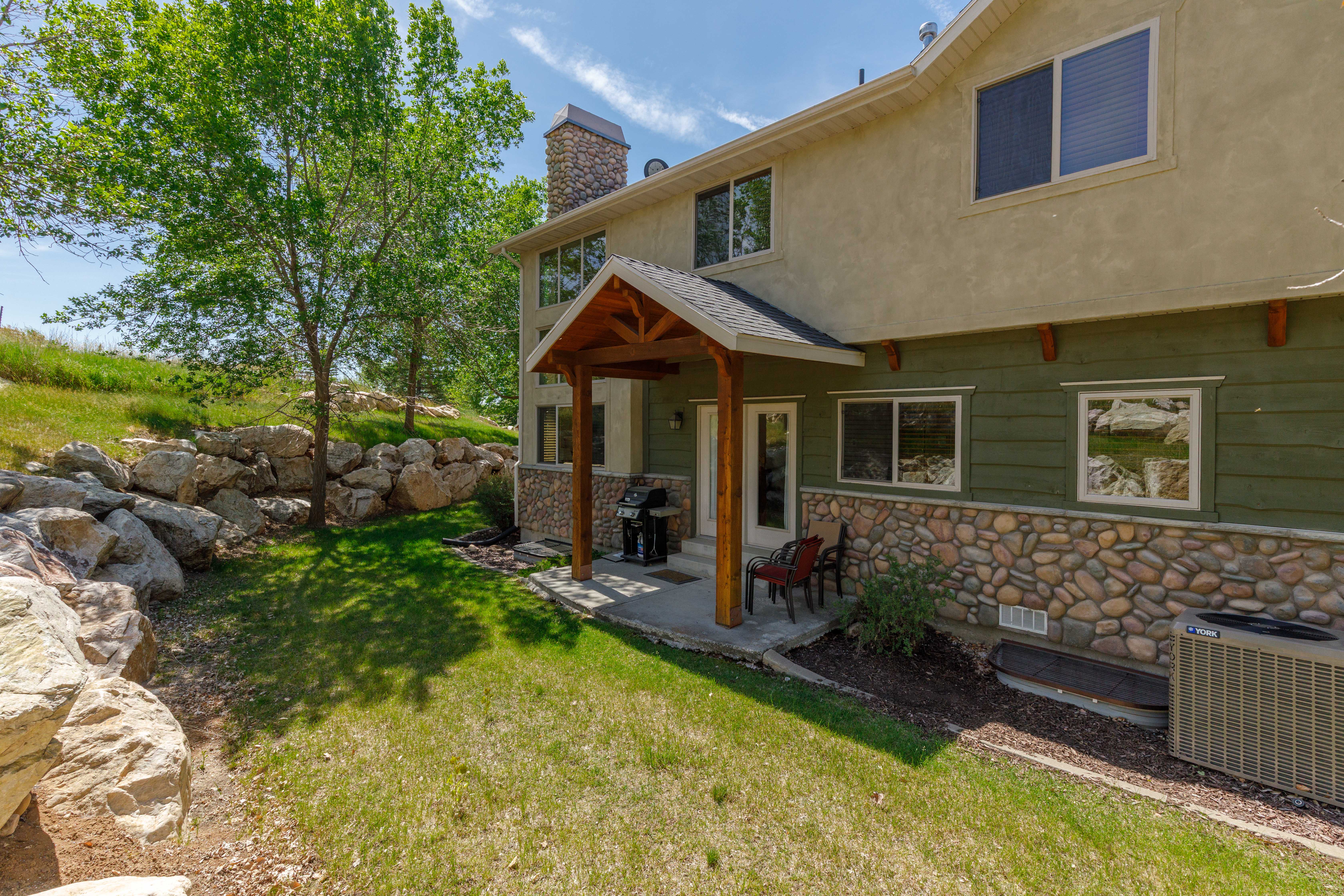 Rear exterior featuring a covered patio with exposed wood beams, a stone-clad foundation, a stucco upper level, and a sloped lawn with large rock landscaping