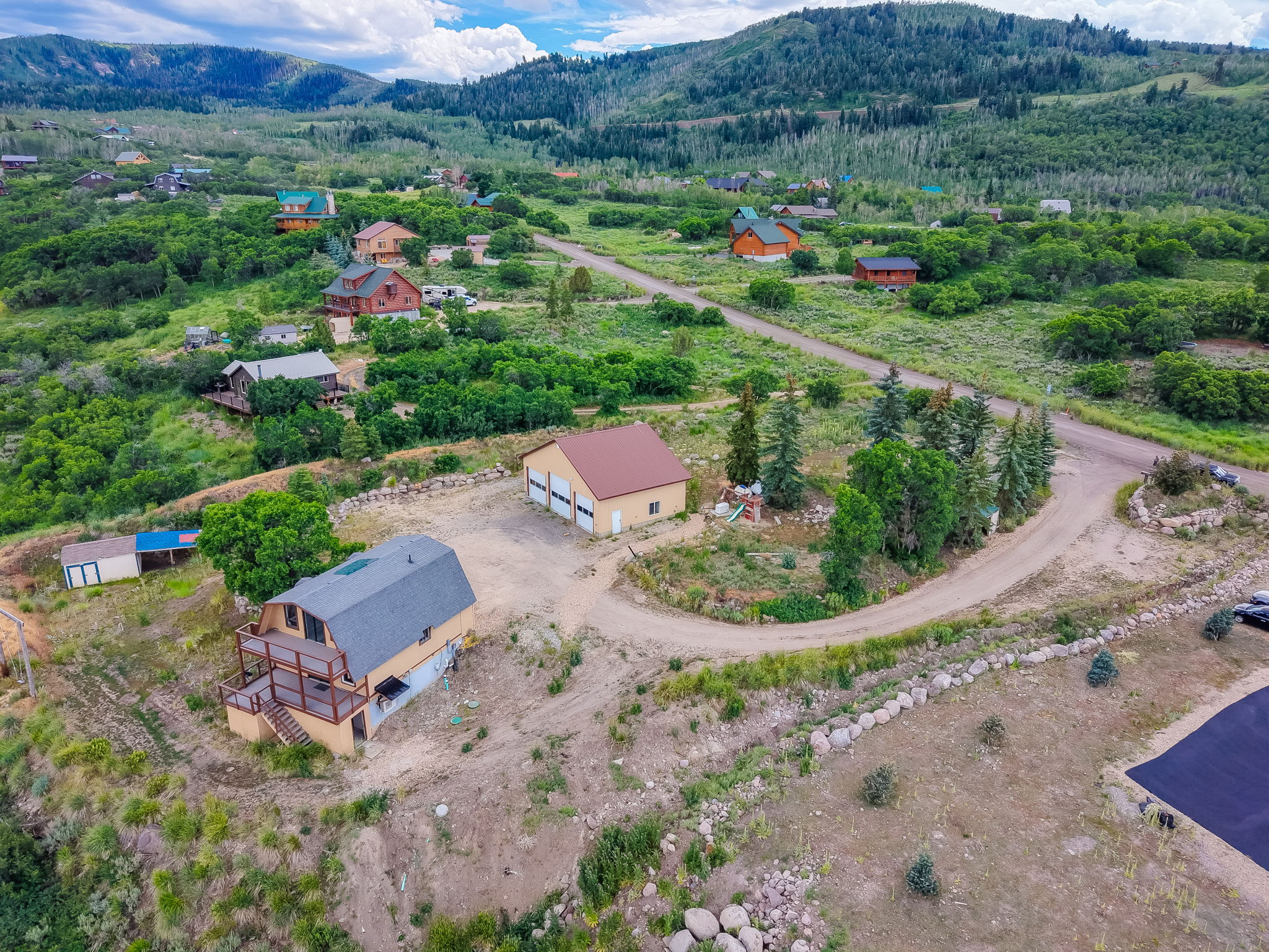 Expansive property featuring a multi-level deck, detached garage with multiple bays, secondary shed, dirt driveway, and surrounding natural foliage