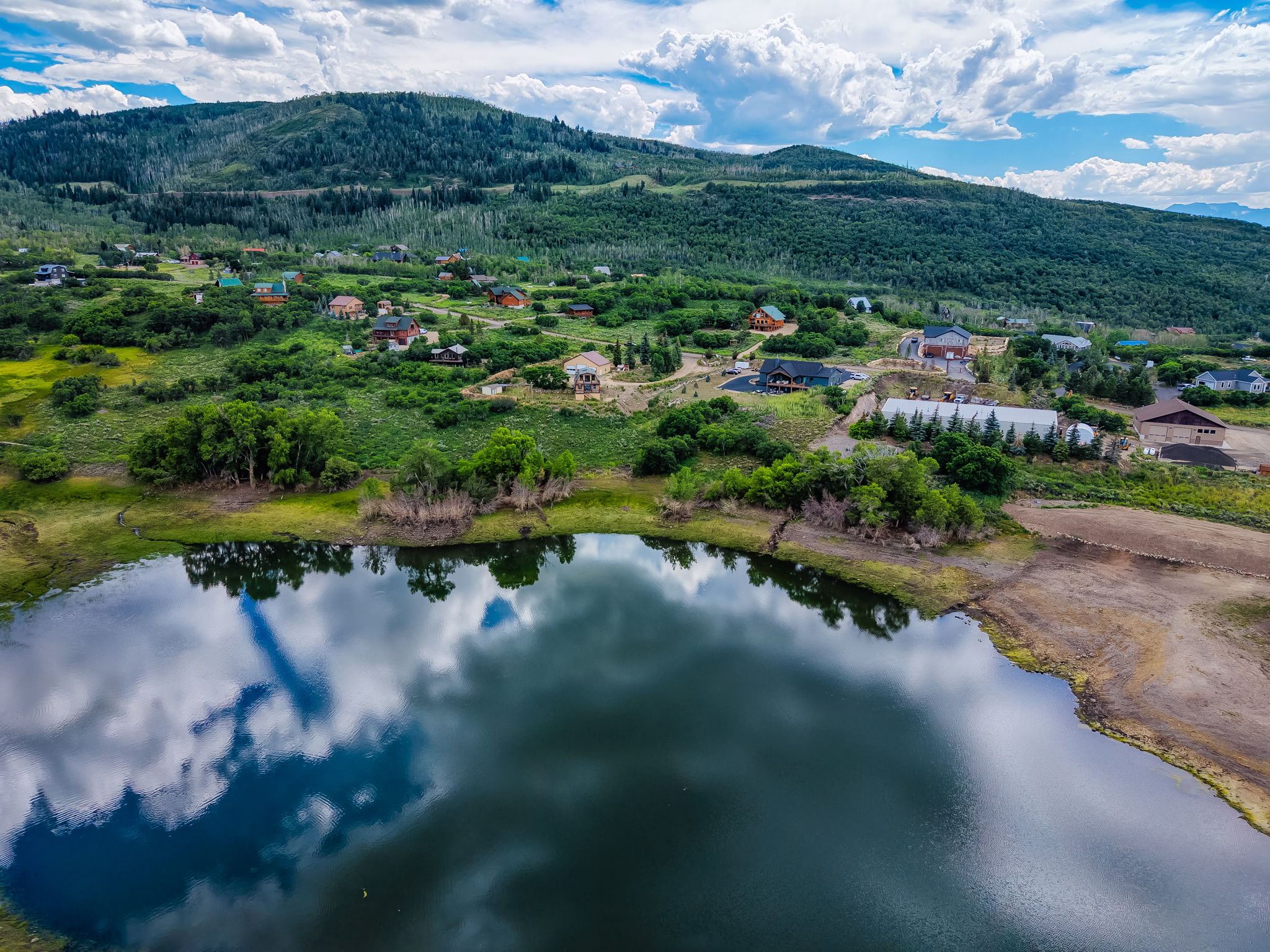 Expansive mountain landscape featuring a reflective body of water, lush green hillsides, scattered residences, and mature tree coverage