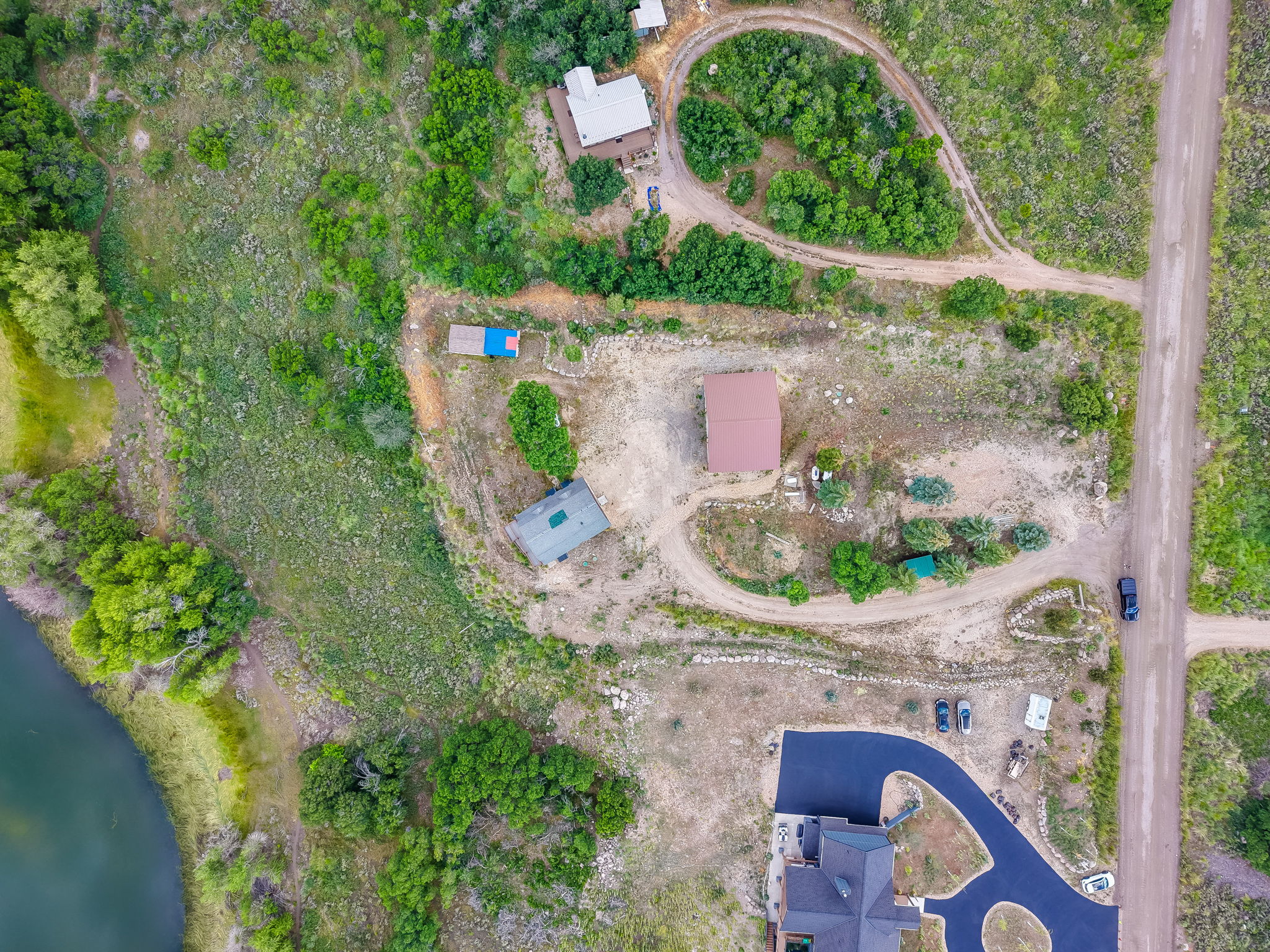 Expansive aerial property showcasing multiple structures, a winding unpaved road, a paved driveway, a body of water, and abundant natural vegetation