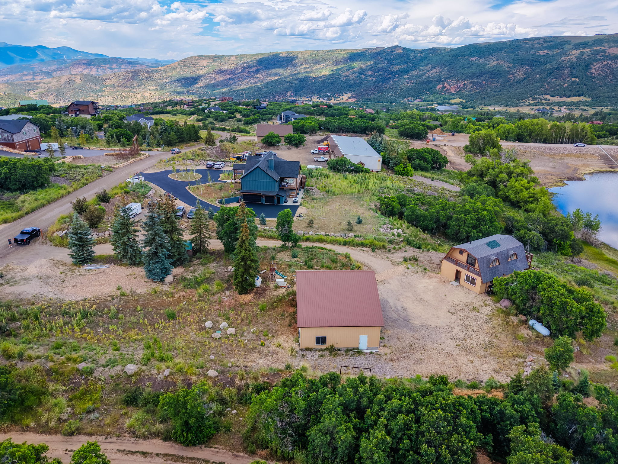 Expansive property featuring a contemporary home with a circular paved driveway, a separate barn-style structure with dormer windows, a detached building with a metal roof, and a large body of water