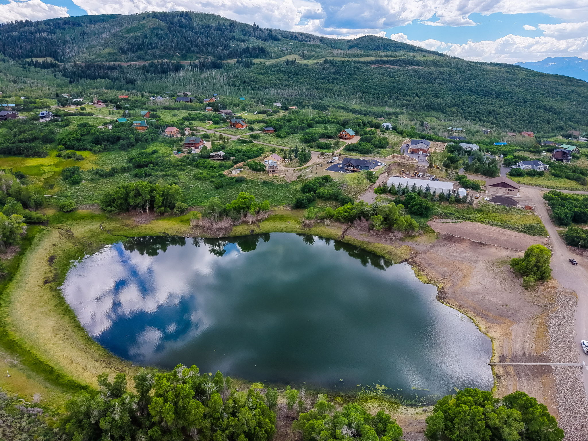 Expansive mountain-backed property featuring a calm lake with cloud reflections, surrounding green foliage, and a collection of residential structures
