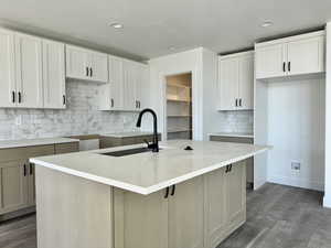 Two tone kitchen featuring tasteful backsplash, dark wood finished floors, a center island with sink, light stone countertops, and recessed lighting