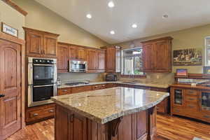 Kitchen with stainless steel appliances, light stone counters, a center island, lofted ceiling, and wood finish cabinets