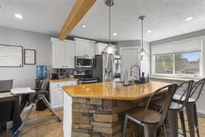 Kitchen featuring white cabinets, stainless steel appliances, hanging light fixtures, a kitchen island with sink, and beam ceiling