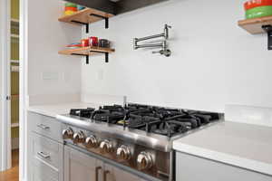 Kitchen view of stainless steel gas cooktop, pot filler, quartz countertops, open shelves, and cabinetry