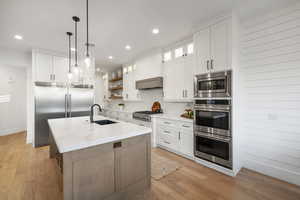 Two tone kitchen featuring glass fronted cabinets, light stone countertops, an island with sink, built in appliances, and open shelves