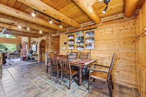 Dining space with track lighting, a wooden ceiling with exposed beams, rustic walls, wooden walls, and stone tile floors