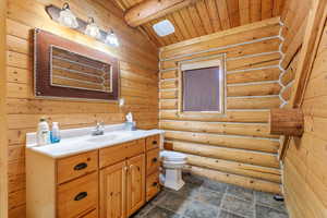 Bathroom with vanity, wooden ceiling, vaulted ceiling with beams, rustic walls, and dark stone finish floors