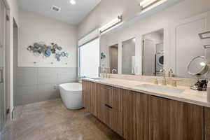 Full bath with double vanity, a freestanding bath, washer / dryer, tile walls, and concrete flooring