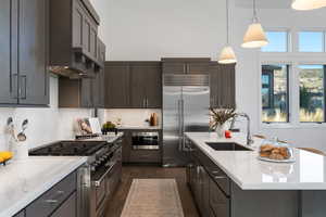 Kitchen with premium appliances, light stone counters, dark wood finish cabinets, and a center island with sink
