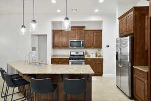 Kitchen with stainless steel appliances, light stone counters, decorative light fixtures, a kitchen breakfast bar, and an island with sink