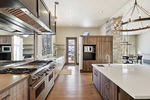 Kitchen with stainless steel appliances, premium range hood, light stone counters, light wood-style floors, and an island with sink