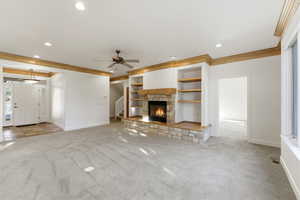 Unfurnished living room featuring light colored carpet, a stone fireplace, ceiling fan, ornamental molding, and recessed lighting