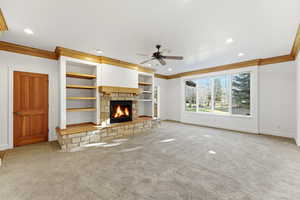 Unfurnished living room featuring ornamental molding, a stone fireplace, ceiling fan, carpet floors, and built in features