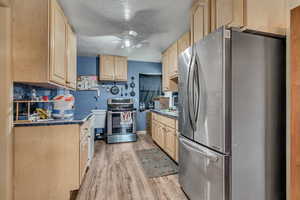 Kitchen featuring light wood finish cabinetry, stainless steel appliances, a textured ceiling, dark countertops, and light wood-type flooring