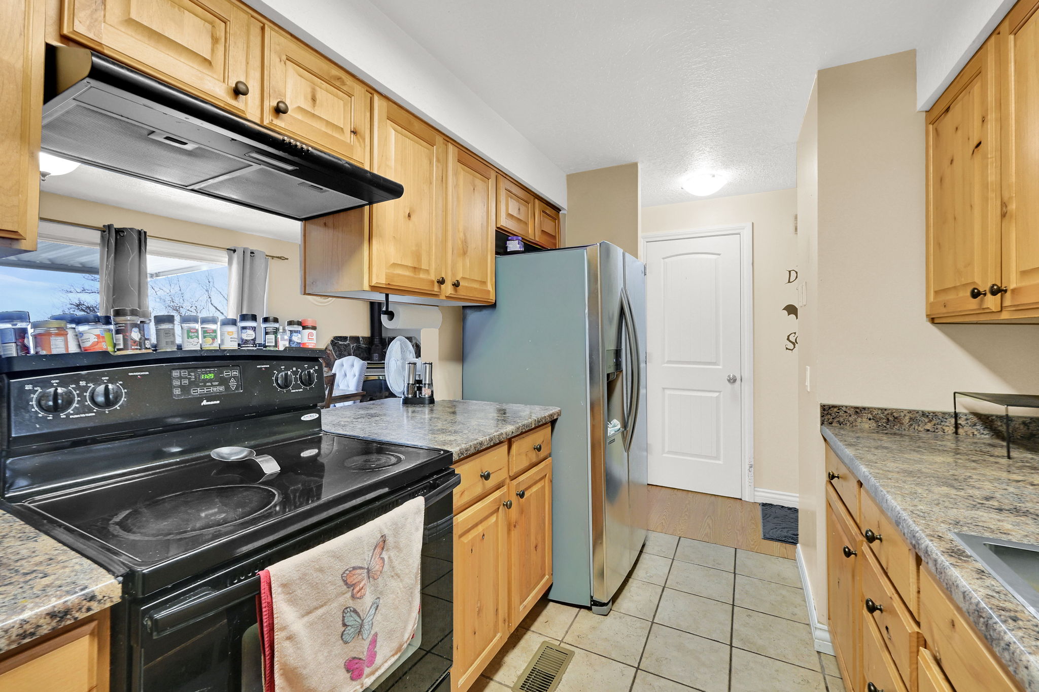 Kitchen with electric range, range hood, stainless steel fridge with ice dispenser, light tile patterned floors, and light wood finish cabinets