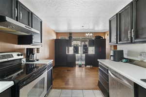 Kitchen with stainless steel appliances, light countertops, a textured ceiling, suspended lighting, and dark cabinetry