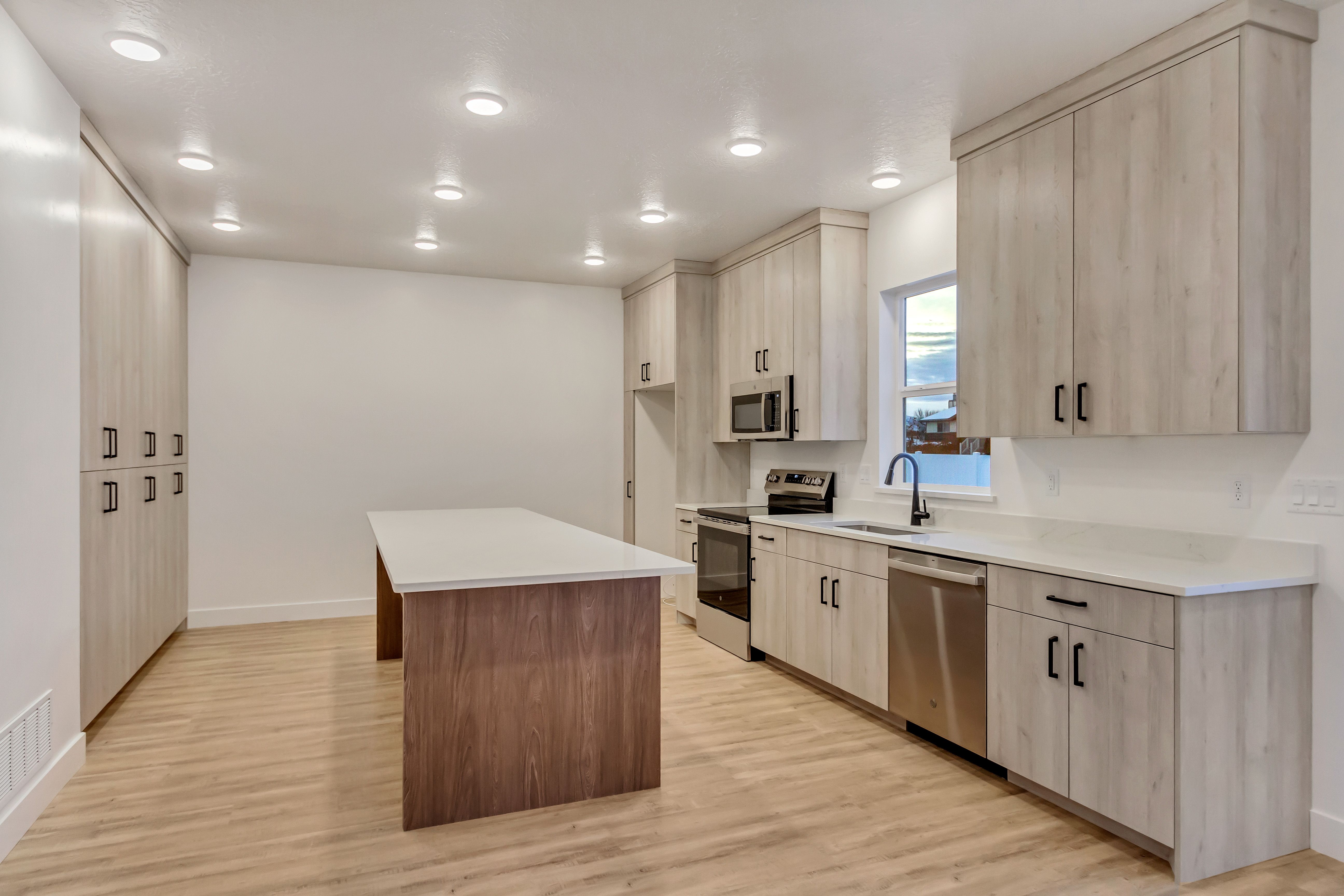 Kitchen featuring an elongated island with a contrasting wood-finish base and light countertop, light wood-finish cabinetry, stainless steel appliances, and wood-finish flooring