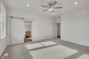 Unfurnished bedroom featuring a barn door, connected bathroom, light colored carpet, recessed lighting, and a ceiling fan