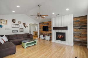 Living area featuring arched walkways, wood walls, light wood-type flooring, a large fireplace, and a ceiling fan
