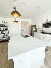 Kitchen with light wood-style flooring, a center island with sink, light stone counters, white cabinetry, and decorative light fixtures
