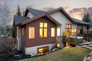 Back of house at dusk featuring a shingled roof, a deck, a lawn, stucco siding, and stone siding