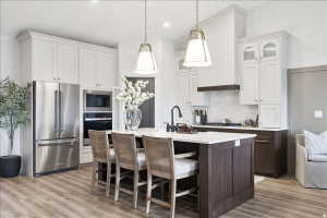 Modern kitchen featuring a central island with white countertop, wood-finish flooring, two-tone cabinetry with light upper and dark lower cabinets, stainless steel appliances, and two pendant light fixtures