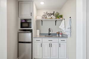Bar with stainless steel fridge, white cabinets, open shelves, black microwave, and light stone countertops