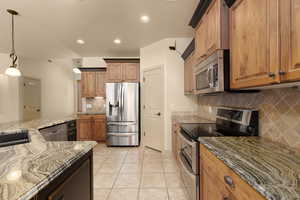 Kitchen featuring stainless steel appliances, dark stone counters, light tile patterned floors, wood finish cabinetry, and decorative light fixtures