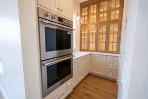 Kitchen with white cabinets, stainless steel double oven, dark wood-style floors, and glass fronted cabinets