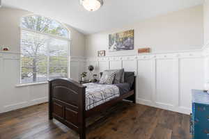Bedroom featuring a decorative wall, dark wood-style flooring, wainscoting, and vaulted ceiling