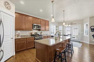 Kitchen featuring wood finish cabinetry, dark wood flooring, stainless steel appliances, light stone countertops, and an island with sink