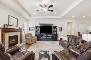 Living room featuring a tile fireplace, wood finished floors, a raised ceiling, ceiling fan, and arched walkways