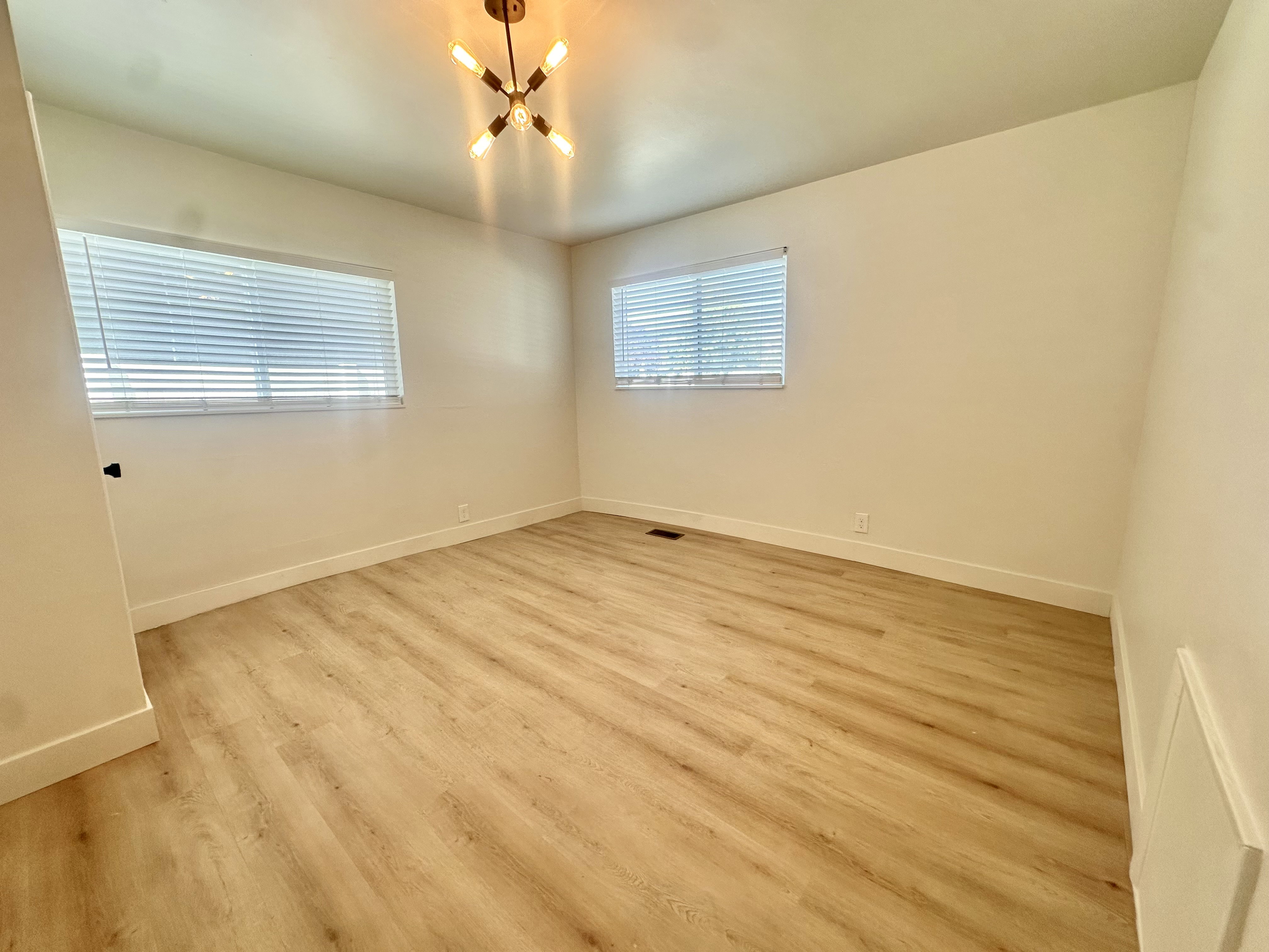 Spacious room with wood-finish flooring, two windows with blinds, a modern ceiling light fixture, and white baseboards